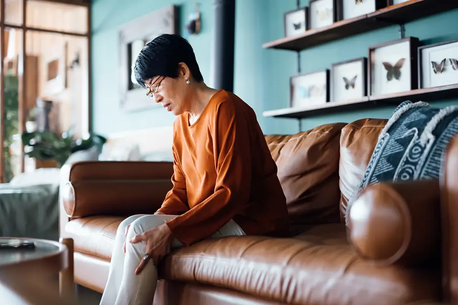 Senior woman sitting on a couch holding her knee with both hands, indicating knee pain or joint discomfort.