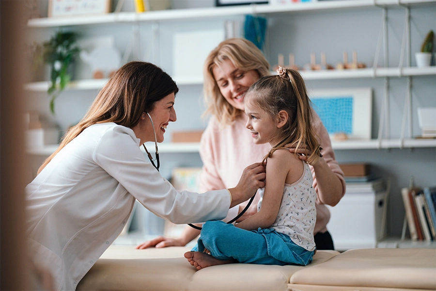Smiling pediatrician listens to a young girl's heartbeat with a stethoscope while her mother holds her during a checkup.